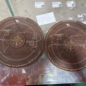 May include: Two circular dark brown wooden coin banks featuring an engraved world map, a central compass design with the text Adventure Awaits, and the phrase Coins may fill your pocket but adventure will fill your soul along the edge.