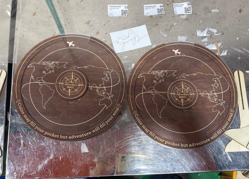 May include: Two circular dark brown wooden coin banks featuring an engraved world map, a central compass design with the text Adventure Awaits, and the phrase Coins may fill your pocket but adventure will fill your soul along the edge.