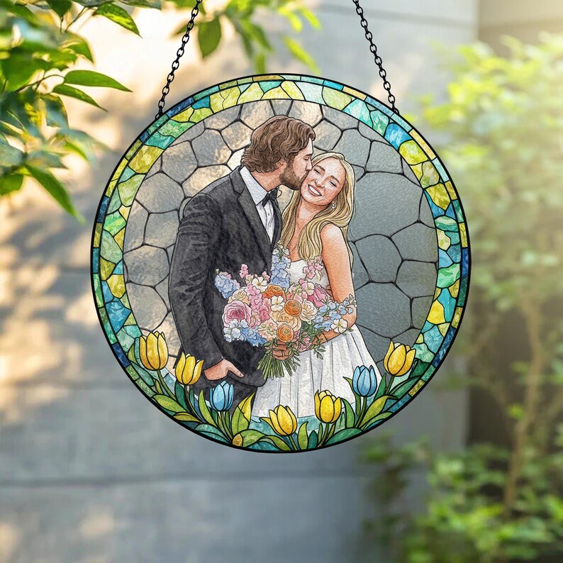 May include: A round stained glass window featuring a couple embracing, with the man kissing the woman's cheek. The artwork is surrounded by a border of colorful glass and floral accents, including yellow and blue tulips. The piece hangs from a black chain.