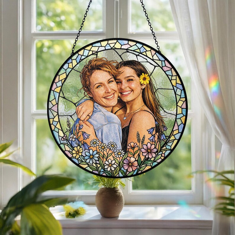 May include: A round stained glass window with a black metal chain for hanging. The window features a portrait of two women embracing, surrounded by a floral border with daisies and bluebells. The glass has a colorful, mosaic-like design.