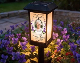 May include: A black solar-powered memorial lantern with a rectangular glass panel featuring a portrait of a person, purple floral illustrations, and the text "FOREVER IN OUR HEARTS", "A BEAUTIFUL SOUL NEVER FORGOTTEN", "Evelyn Grace", and "1941 - 2024".