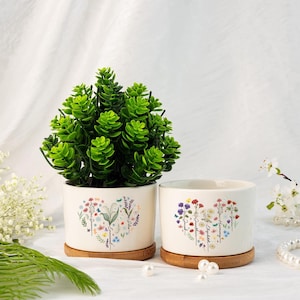 Könnte beinhalten: Zwei weiße Keramik-Blumentöpfe mit einem herzförmigen Design und Blumenmustern. Die Töpfe tragen die Namen "Grandma", "Esther", "Samantha" und "Gwen". Die Töpfe stehen auf einer weißen Oberfläche mit einer grünen Pflanze im linken Topf.