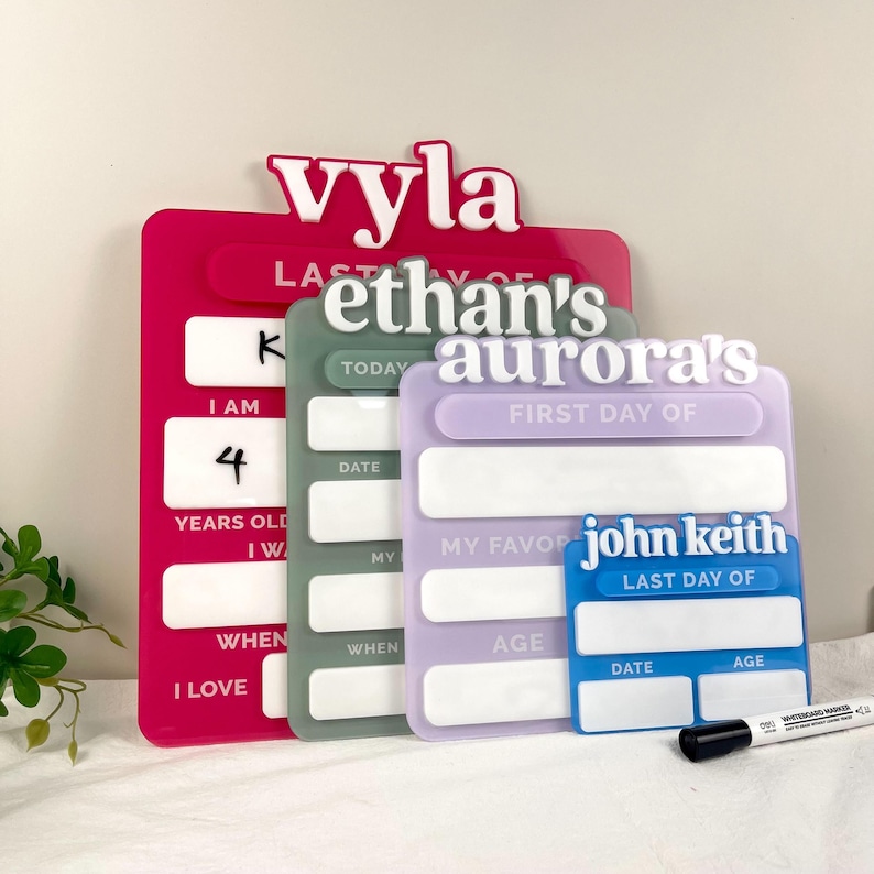May include: Four rectangular acrylic signs in pink, sage green, lavender, and blue, each featuring a raised white name at the top and white text for tracking school milestones. The signs include blank white spaces for writing with the included marker.