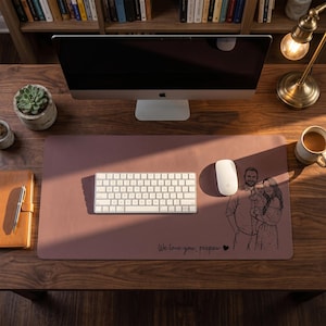 Peut inclure: Un tapis de bureau mauve avec un clavier blanc, une souris et une illustration de portrait de famille. Le texte "We love you, peepaw" est imprim&eacute; sur le tapis. Un &eacute;cran d'ordinateur, une tasse de caf&eacute; et une plante succulente sont &eacute;galement visibles.