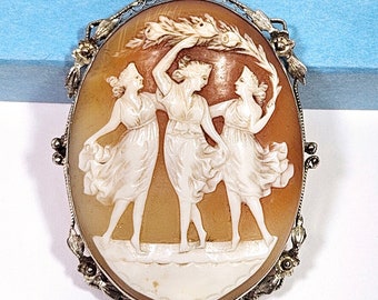 May include: An oval cameo brooch featuring three women in white dresses holding a wreath of leaves. The cameo is set in a silver frame with gold accents.