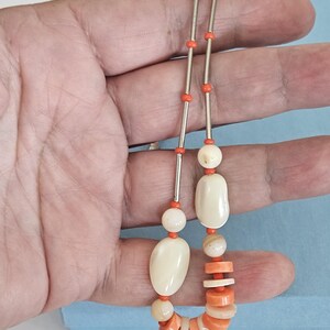 May include: A necklace featuring a combination of bright orange cylindrical beads, small orange round beads, and larger smooth off-white oval beads. The design incorporates alternating segments of metallic silver-toned tubes and orange beads.