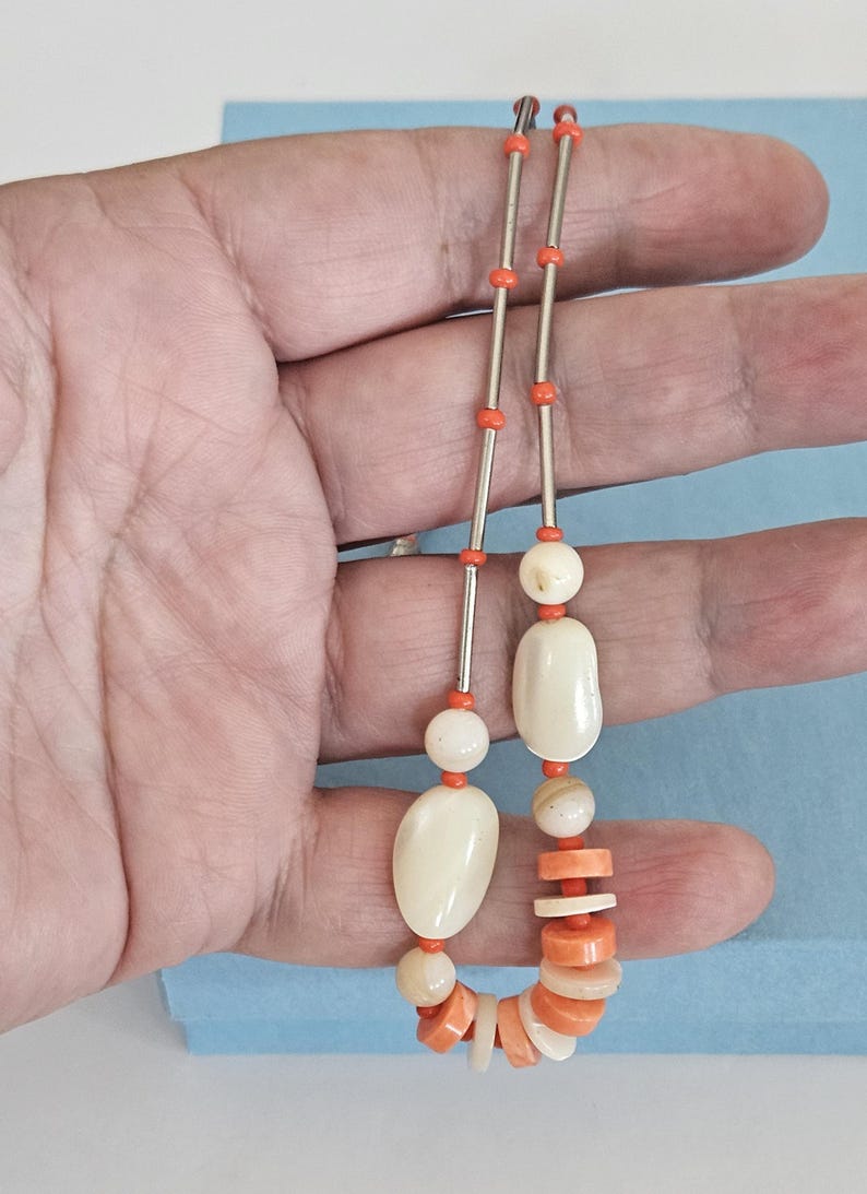 May include: A necklace featuring a combination of bright orange cylindrical beads, small orange round beads, and larger smooth off-white oval beads. The design incorporates alternating segments of metallic silver-toned tubes and orange beads.