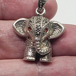 May include: A silver elephant pendant with marcasite detailing and red gemstone eyes. The pendant has a textured surface and a loop for attaching to a chain. The elephant's ears are large and detailed, and the trunk is curled.