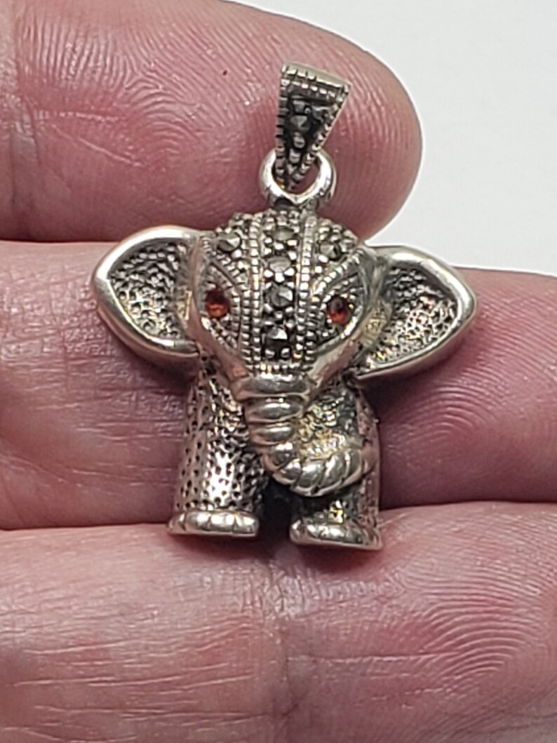 May include: A silver elephant pendant with marcasite detailing and red gemstone eyes. The pendant has a textured surface and a loop for attaching to a chain. The elephant's ears are large and detailed, and the trunk is curled.
