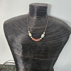 May include: A necklace featuring a series of small, cylindrical coral-colored beads interspersed with larger, smooth, off-white oval stones. The beads are strung on a thin, light-colored cord, creating a delicate arrangement that centers on the neck.