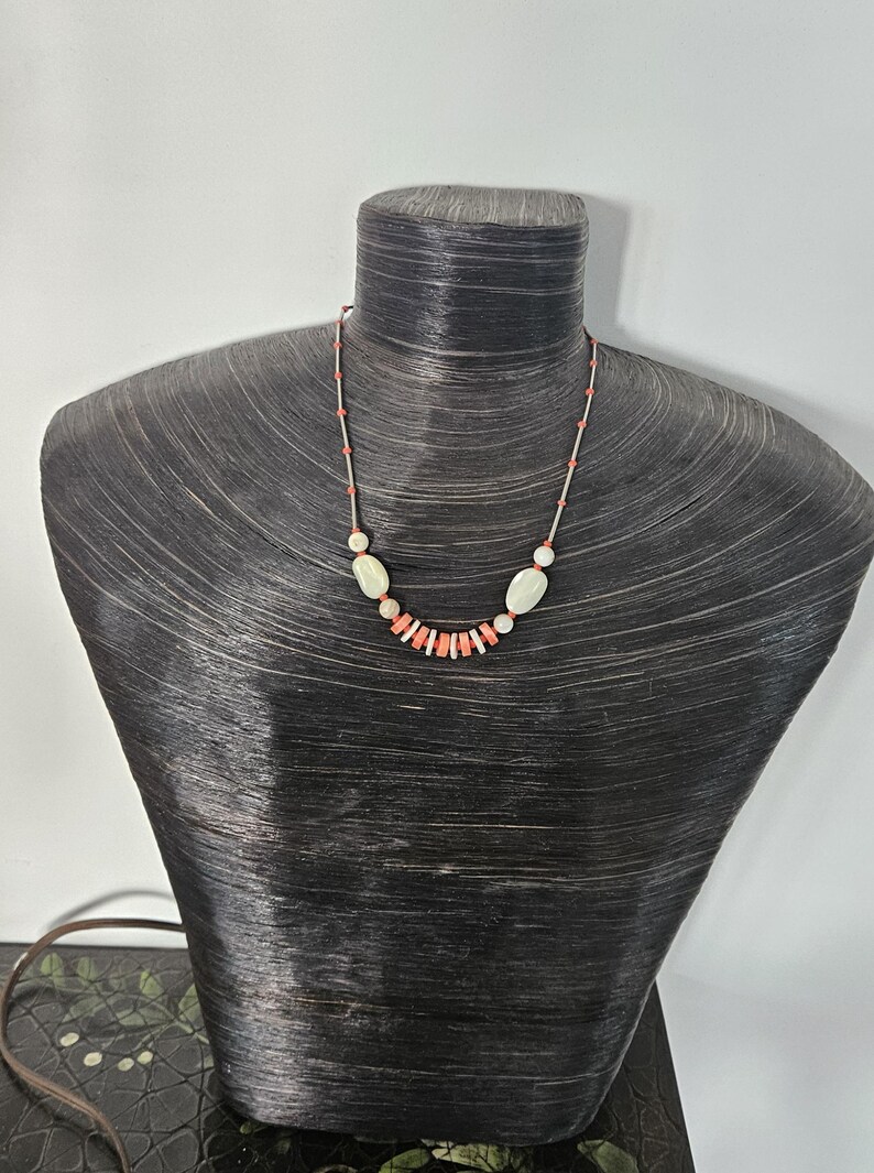 May include: A necklace featuring a series of small, cylindrical coral-colored beads interspersed with larger, smooth, off-white oval stones. The beads are strung on a thin, light-colored cord, creating a delicate arrangement that centers on the neck.