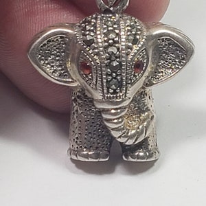 May include: A silver elephant pendant with marcasite detailing. The elephant has red gemstone eyes and a textured surface. The pendant is suspended from a silver bail, showcasing a detailed, miniature animal figurine.