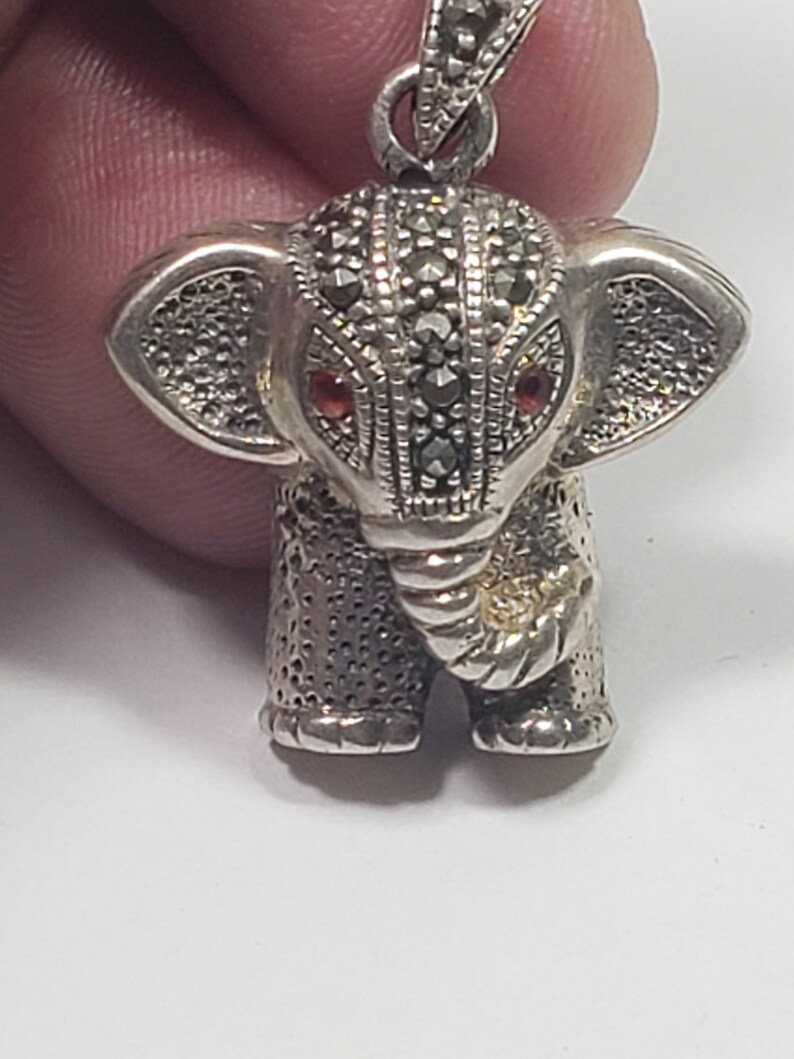 May include: A silver elephant pendant with marcasite detailing. The elephant has red gemstone eyes and a textured surface. The pendant is suspended from a silver bail, showcasing a detailed, miniature animal figurine.