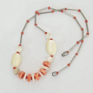 May include: A necklace featuring a sequence of silver-toned metal tubes interspersed with small orange beads. The central section includes larger, smooth, cream-colored oval beads and a series of alternating flat, circular orange and white discs.