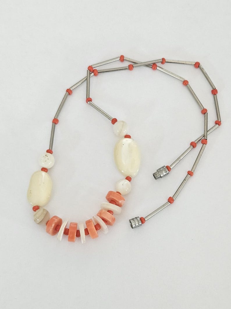 May include: A necklace featuring a sequence of silver-toned metal tubes interspersed with small orange beads. The central section includes larger, smooth, cream-colored oval beads and a series of alternating flat, circular orange and white discs.