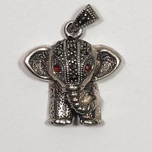 May include: A silver elephant pendant with black and red accents. The elephant has detailed markings and small red eyes. The pendant has a loop at the top for a chain. The elephant's head and body are covered in small, dark stones.
