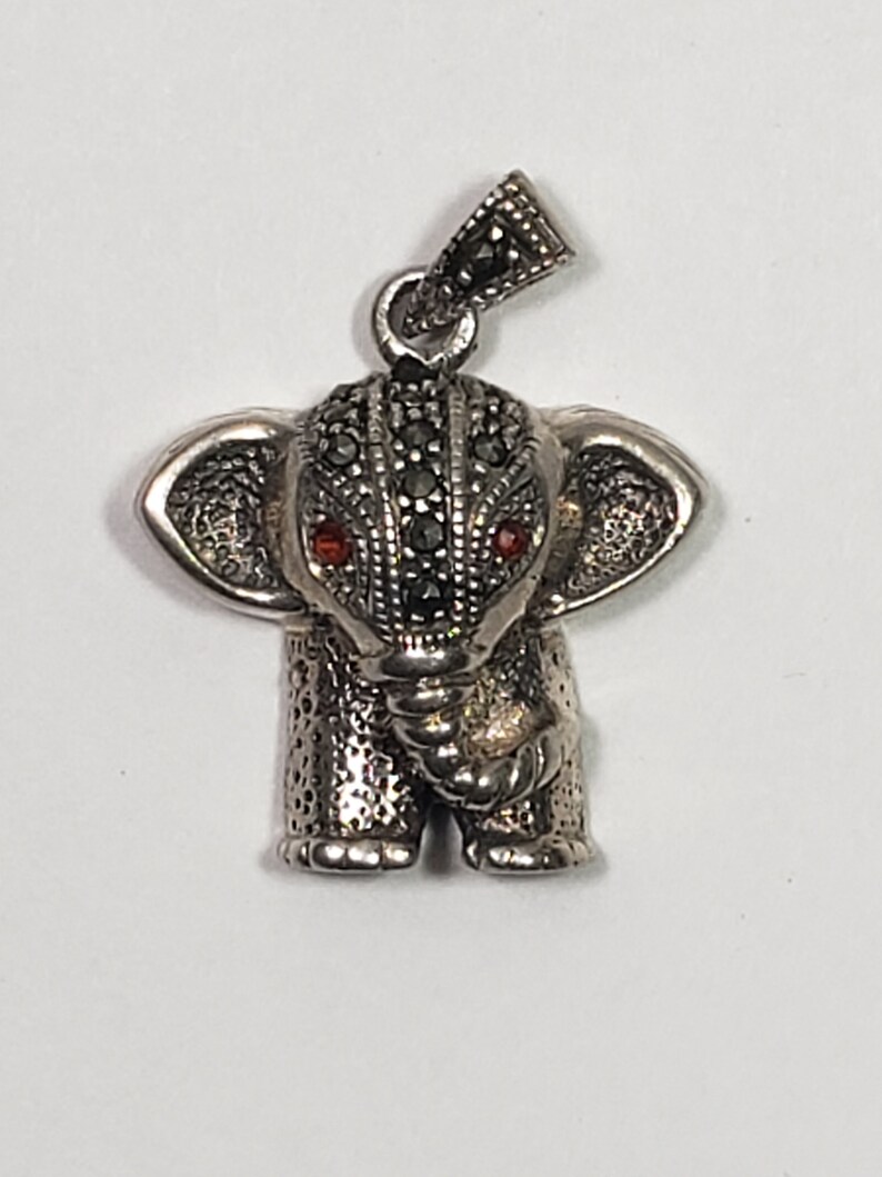 May include: A silver elephant pendant with black and red accents. The elephant has detailed markings and small red eyes. The pendant has a loop at the top for a chain. The elephant's head and body are covered in small, dark stones.
