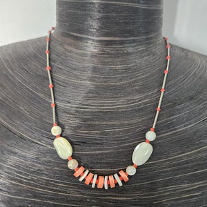 May include: A necklace featuring a series of thin silver-toned metal tubes interspersed with small coral-colored beads. The center section includes two smooth, pale oval beads and a row of alternating flat, disc-shaped coral and white beads.