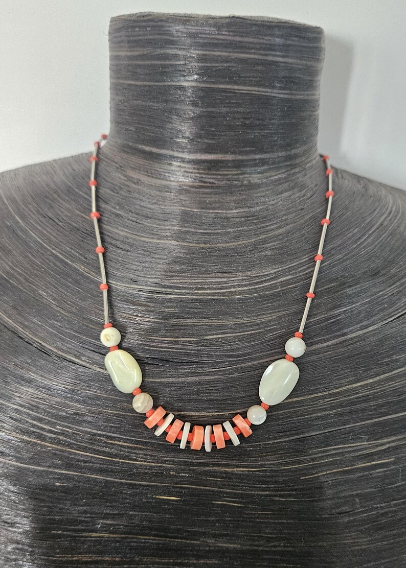 May include: A necklace featuring a series of thin silver-toned metal tubes interspersed with small coral-colored beads. The center section includes two smooth, pale oval beads and a row of alternating flat, disc-shaped coral and white beads.