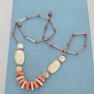 May include: A necklace featuring a series of silver-toned metal tubes interspersed with small orange beads. The lower section includes larger, smooth, off-white oval beads and a cluster of flat, circular orange and white beads arranged in a row.