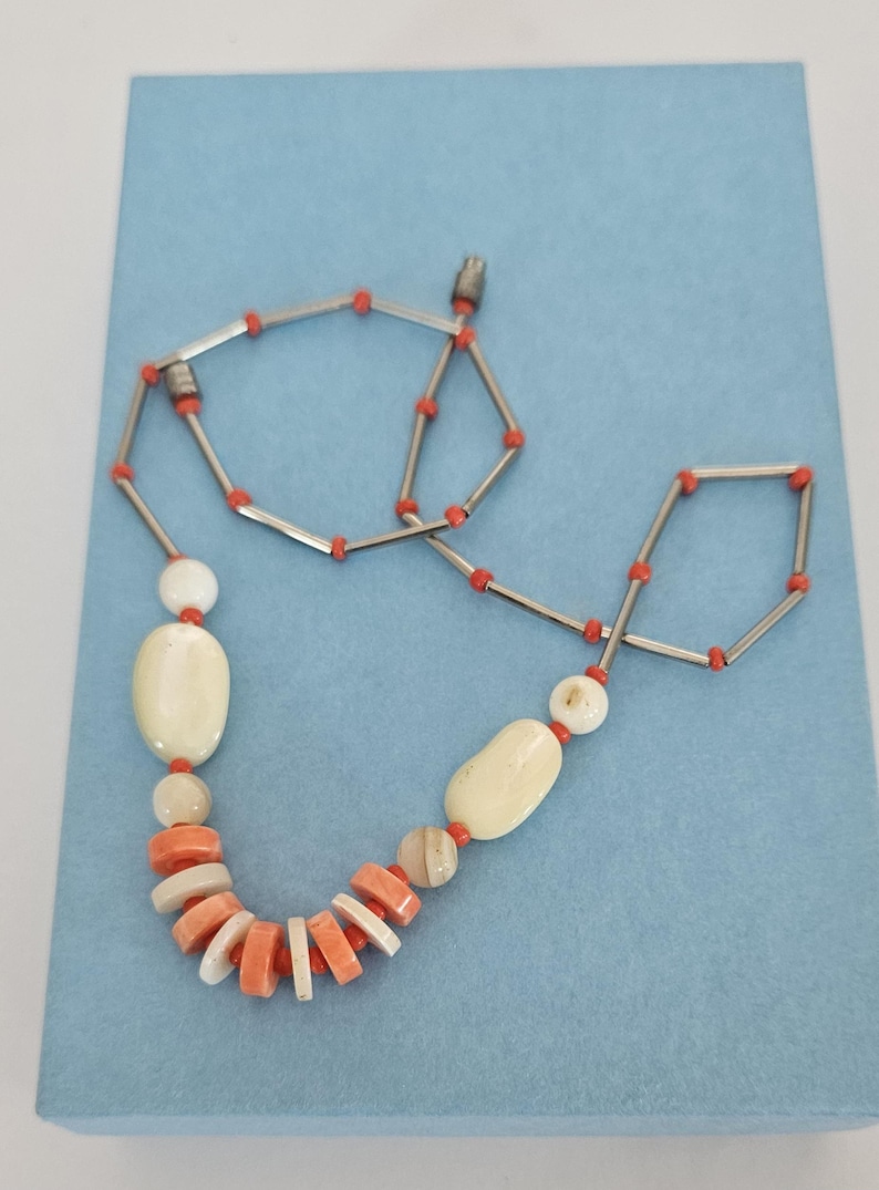 May include: A necklace featuring a series of silver-toned metal tubes interspersed with small orange beads. The lower section includes larger, smooth, off-white oval beads and a cluster of flat, circular orange and white beads arranged in a row.