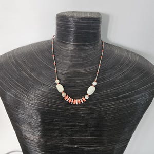 May include: A necklace featuring a series of small coral-colored cylindrical beads and larger smooth white oval beads strung on a thin silver-toned wire. The design alternates between the coral and white elements, creating a balanced pattern.