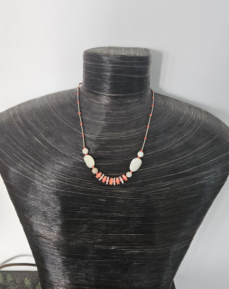 May include: A necklace featuring a series of small coral-colored cylindrical beads and larger smooth white oval beads strung on a thin silver-toned wire. The design alternates between the coral and white elements, creating a balanced pattern.