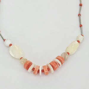 May include: A necklace featuring a strand of thin silver-toned metal tubes interspersed with small bright orange beads. The center section includes larger white oval beads, translucent beige beads, and a series of alternating flat orange and white discs.