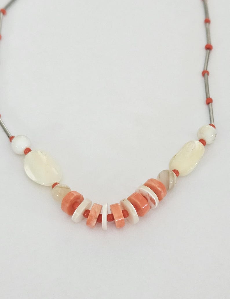 May include: A necklace featuring a strand of thin silver-toned metal tubes interspersed with small bright orange beads. The center section includes larger white oval beads, translucent beige beads, and a series of alternating flat orange and white discs.