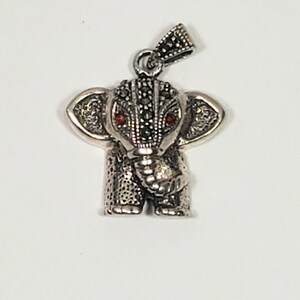 May include: A silver-toned elephant pendant with red gemstone eyes. The pendant has detailed texturing and a small loop for a chain. The elephant's head and ears are adorned with small, dark stones.