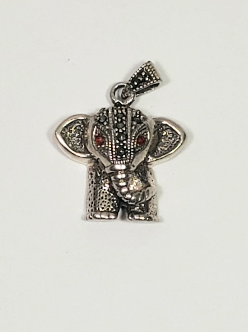 May include: A silver-toned elephant pendant with red gemstone eyes. The pendant has detailed texturing and a small loop for a chain. The elephant's head and ears are adorned with small, dark stones.