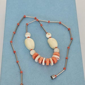 May include: A necklace featuring a series of silver-toned metal tubes interspersed with small orange beads. The center section includes larger, smooth, off-white oval beads and a row of alternating flat, circular orange and white disc beads.