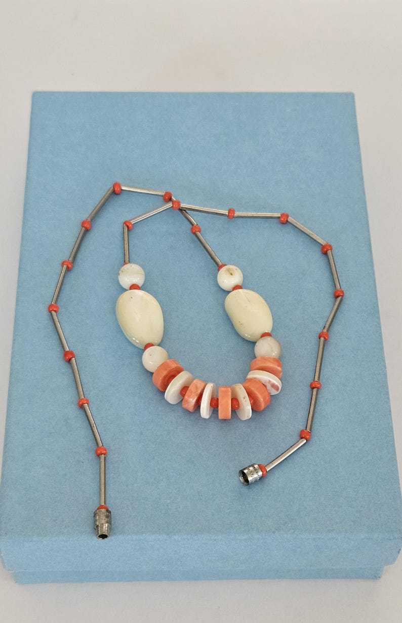 May include: A necklace featuring a series of silver-toned metal tubes interspersed with small orange beads. The center section includes larger, smooth, off-white oval beads and a row of alternating flat, circular orange and white disc beads.