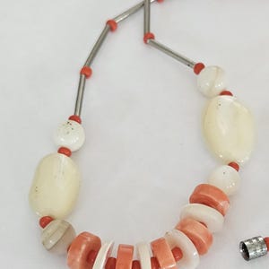 May include: A necklace featuring a sequence of smooth, off-white oval beads, smaller round white beads, and alternating flat, circular coral-colored and white disc beads. The strand is accented with thin silver-toned metal tubes and tiny red beads.