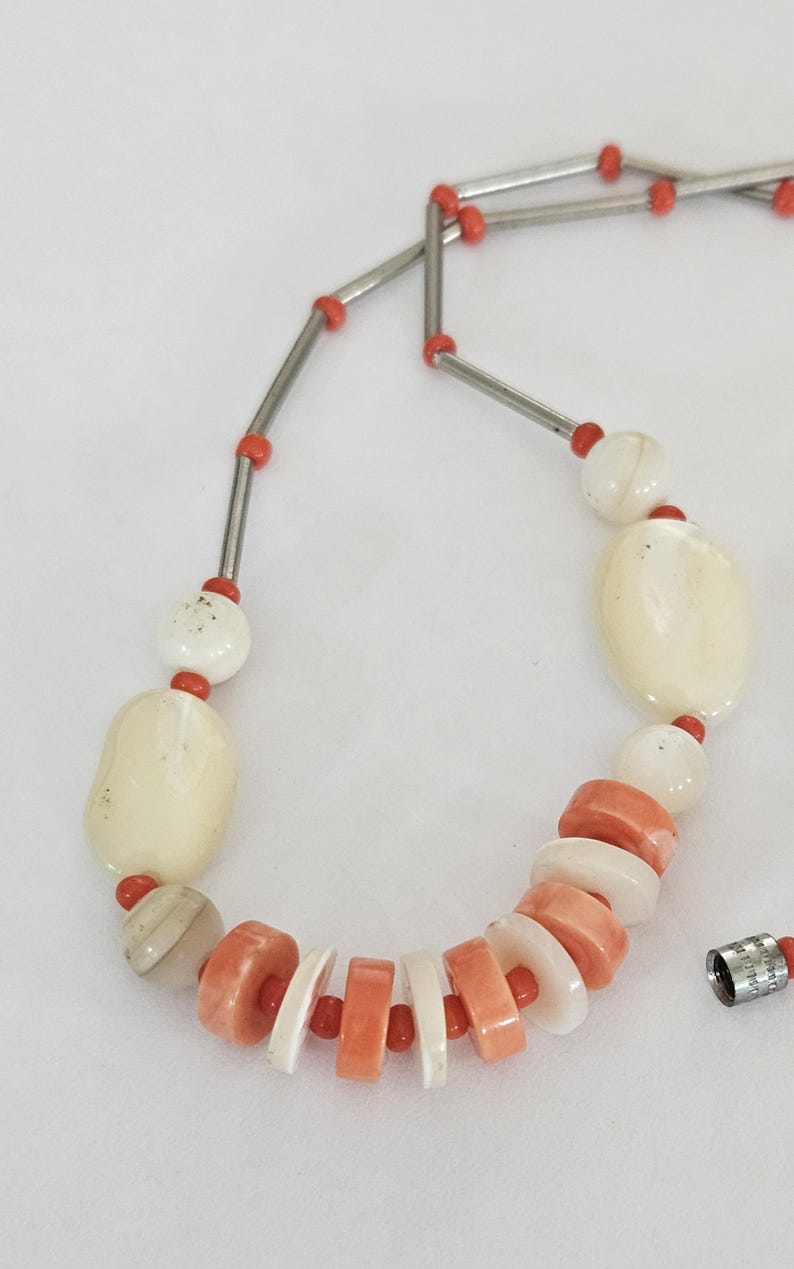 May include: A necklace featuring a sequence of smooth, off-white oval beads, smaller round white beads, and alternating flat, circular coral-colored and white disc beads. The strand is accented with thin silver-toned metal tubes and tiny red beads.