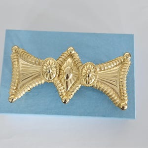 May include: A gold-tone metal hair barrette featuring a wide, elongated shape with a central diamond-shaped motif flanked by two oval accents. The surface displays textured, radiating lines and beaded edges, creating a symmetrical, decorative design.