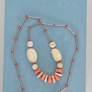 May include: A necklace featuring a long silver-tone chain interspersed with small orange beads. The center section includes two large oval off-white beads, two smaller round off-white beads, and a series of alternating flat orange and white disc beads.
