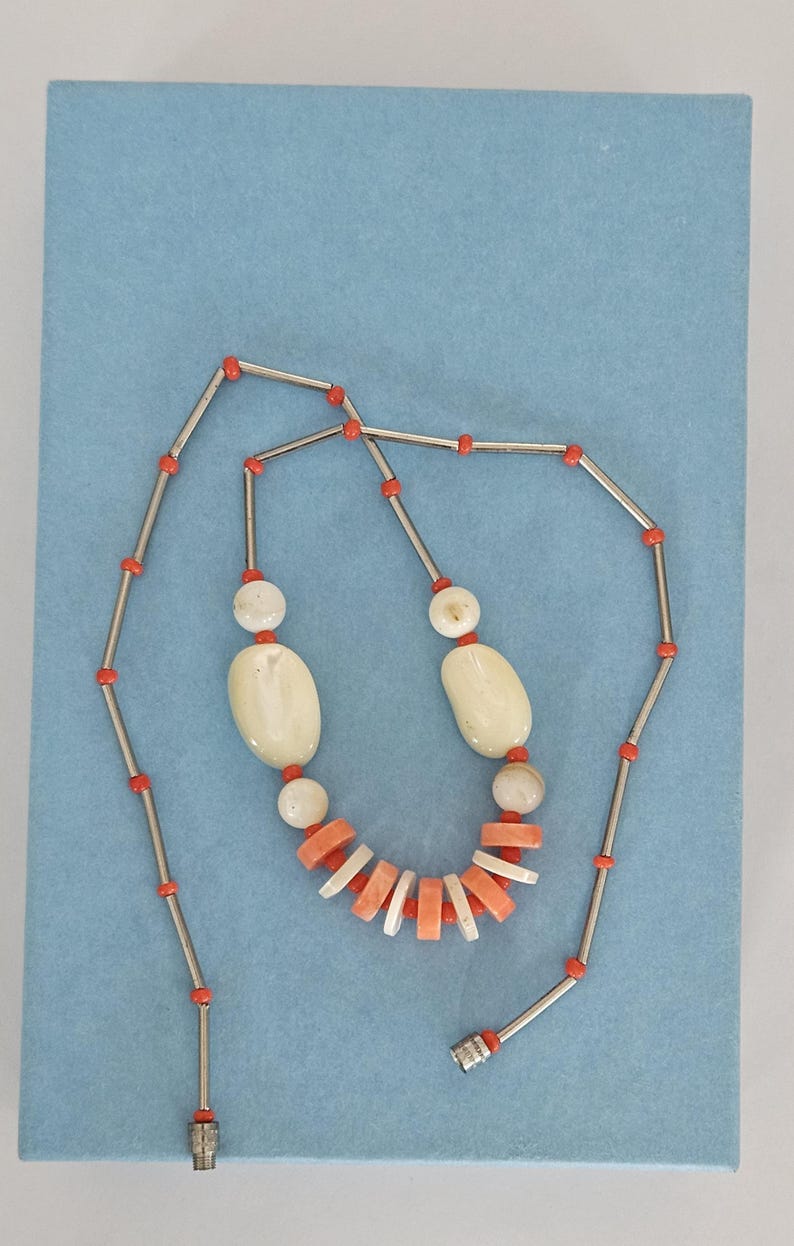 May include: A necklace featuring a long silver-tone chain interspersed with small orange beads. The center section includes two large oval off-white beads, two smaller round off-white beads, and a series of alternating flat orange and white disc beads.
