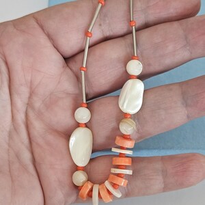 May include: A necklace featuring a mix of orange and white beads. The design includes long silver-toned metal tubes, round white beads, irregular white oval shapes, and flat, circular orange and white disc beads strung on a thin cord.