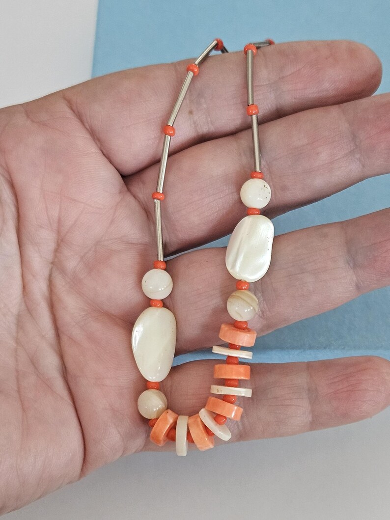 May include: A necklace featuring a mix of orange and white beads. The design includes long silver-toned metal tubes, round white beads, irregular white oval shapes, and flat, circular orange and white disc beads strung on a thin cord.
