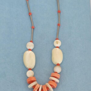 May include: A necklace featuring a series of thin silver-toned metal bars interspersed with small, round, bright orange beads. The lower section includes larger, smooth, off-white oval beads and a cluster of flat, circular orange and white discs.