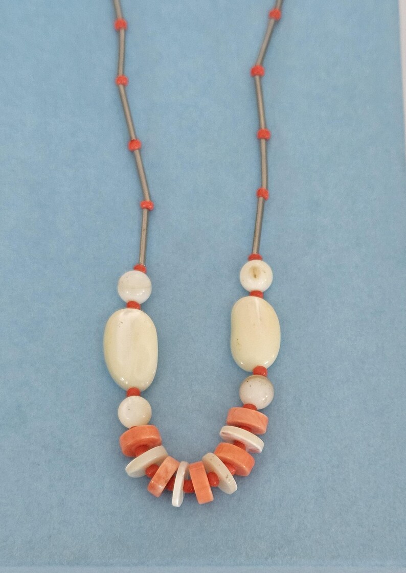 May include: A necklace featuring a series of thin silver-toned metal bars interspersed with small, round, bright orange beads. The lower section includes larger, smooth, off-white oval beads and a cluster of flat, circular orange and white discs.