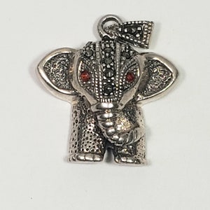 May include: A silver-toned elephant pendant with red gemstone eyes and a textured surface. The pendant has a loop at the top for a chain and is detailed with small, dark stones. The elephant's trunk is curled, and its ears are spread.