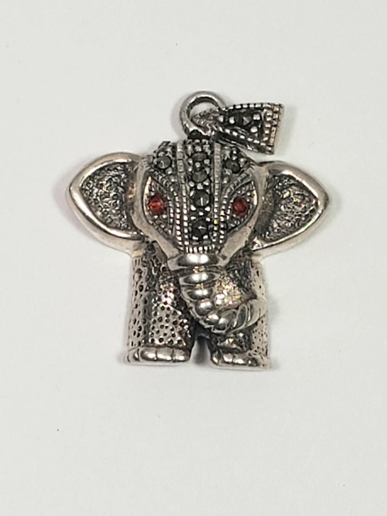 May include: A silver-toned elephant pendant with red gemstone eyes and a textured surface. The pendant has a loop at the top for a chain and is detailed with small, dark stones. The elephant's trunk is curled, and its ears are spread.