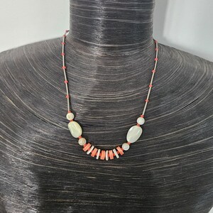 May include: A necklace featuring a thin silver-toned chain interspersed with small orange beads. The center of the necklace displays a series of flat, circular orange and white beads flanked by two larger, smooth, oval-shaped off-white beads.