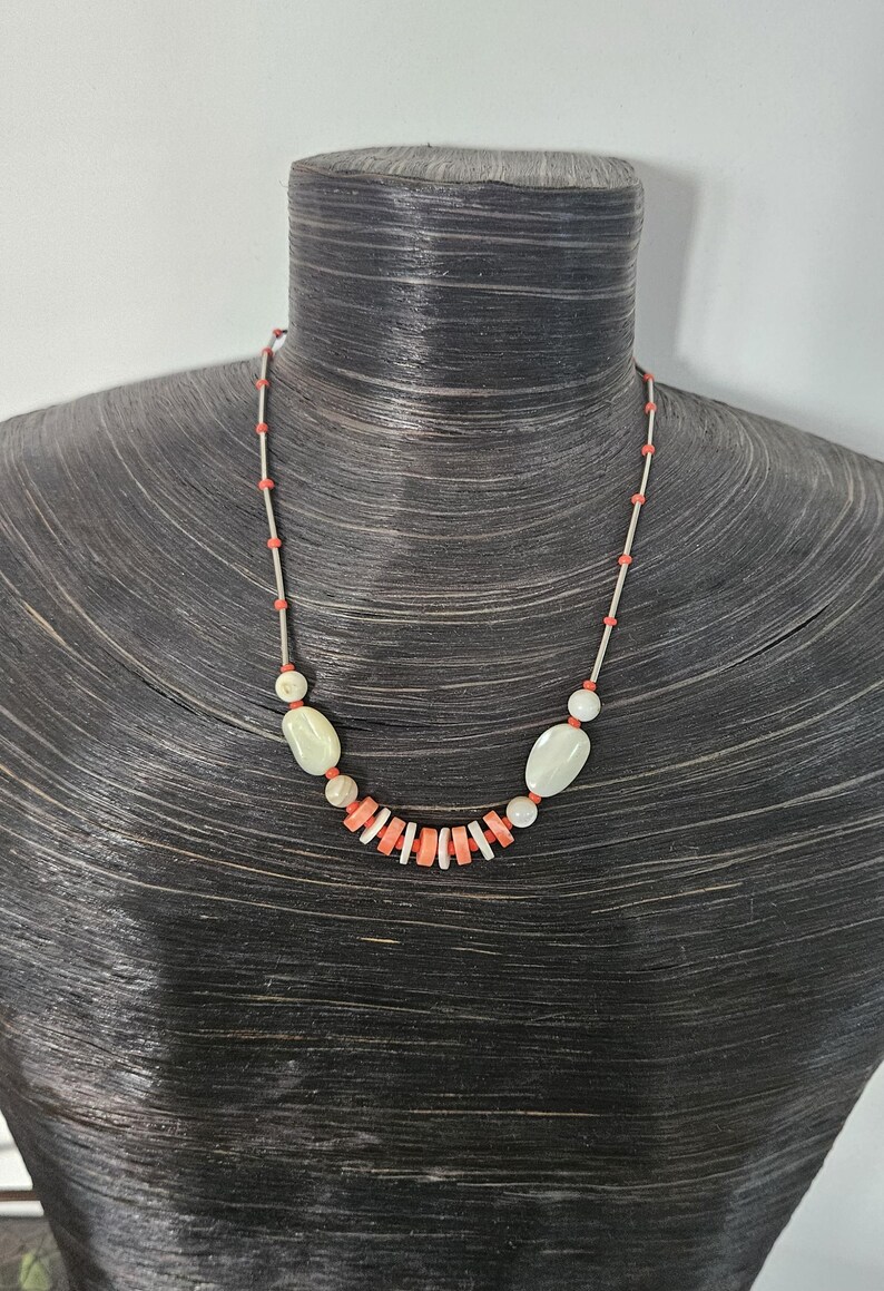 May include: A necklace featuring a thin silver-toned chain interspersed with small orange beads. The center of the necklace displays a series of flat, circular orange and white beads flanked by two larger, smooth, oval-shaped off-white beads.