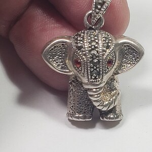 May include: A silver elephant pendant with marcasite detailing and red gemstone eyes. The pendant has a textured surface and a small loop for a chain. The elephant's trunk is curled, and its ears are detailed.