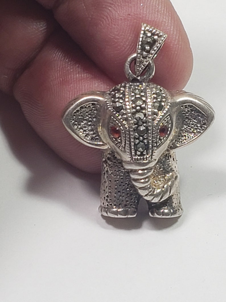 May include: A silver elephant pendant with marcasite detailing and red gemstone eyes. The pendant has a textured surface and a small loop for a chain. The elephant's trunk is curled, and its ears are detailed.