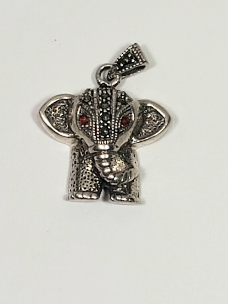 May include: A silver elephant pendant with red gemstone eyes. The pendant has a detailed design, with a textured body and marcasite accents on the head and bail. The elephant's ears are large and detailed.