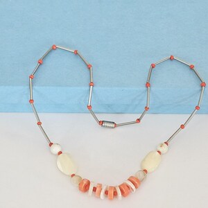 May include: A necklace featuring a series of silver-toned metal tubes separated by small orange beads. The center section includes larger off-white oval beads, translucent round beads, and a cluster of alternating flat orange and white disc beads.